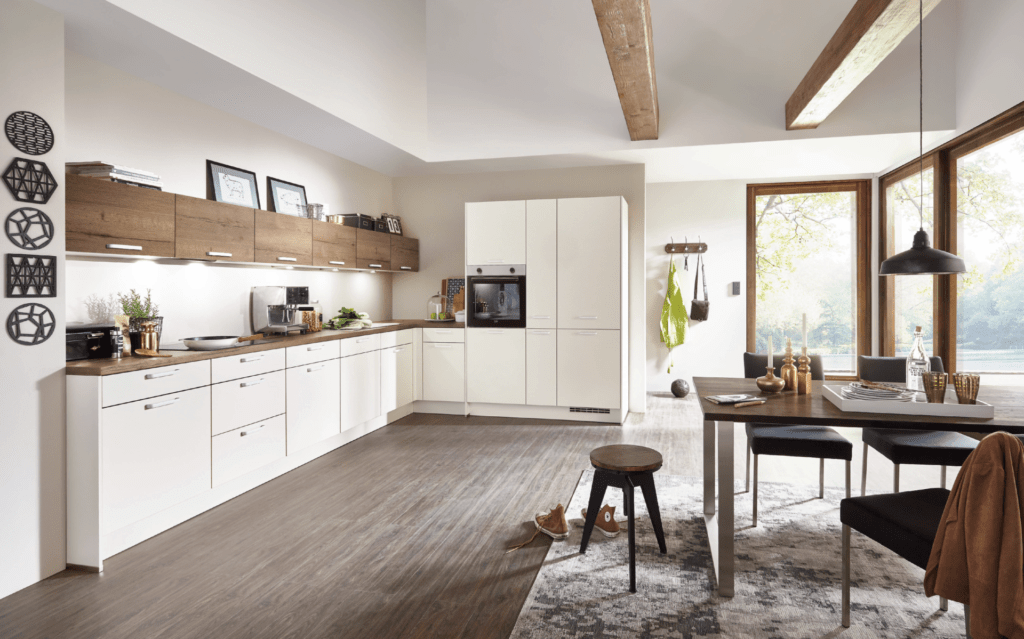 Moderne Nobilia L-Küche in Weiß mit Arbeitsplatte und Oberschränken in warmer Holzoptik, integriertem Backofen und Dunstabzugshaube in einem hellen Raum mit sichtbaren Deckenbalken