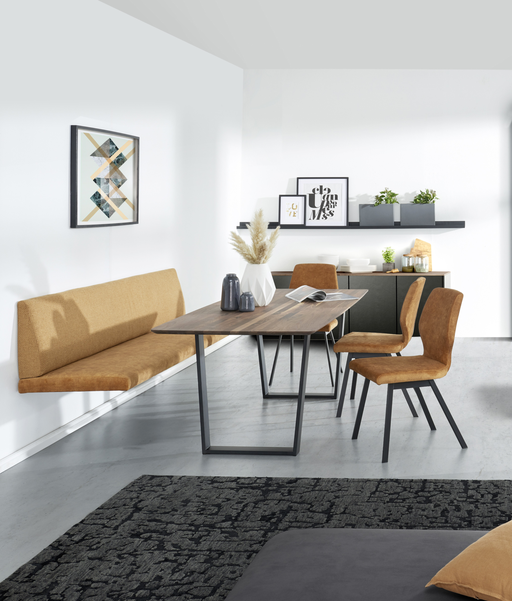 Schösswender Sitzgruppe: gepolsterte Wandbank in Senfgelb mit passendem Esstisch in Dunkeleiche-Optik und schwarzem Trapezgestell, zwei cognacfarbene Veloursstühle – lässiges Esszimmer-Setup vor weißer Wand mit dunklem Sideboard und Wandregal